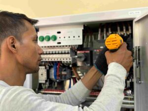 A person uses a yellow cordless drill to work on electrical components inside an open electrical