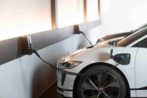 A white electric car is parked indoors, plugged into a wall-mounted charging station—a sign of