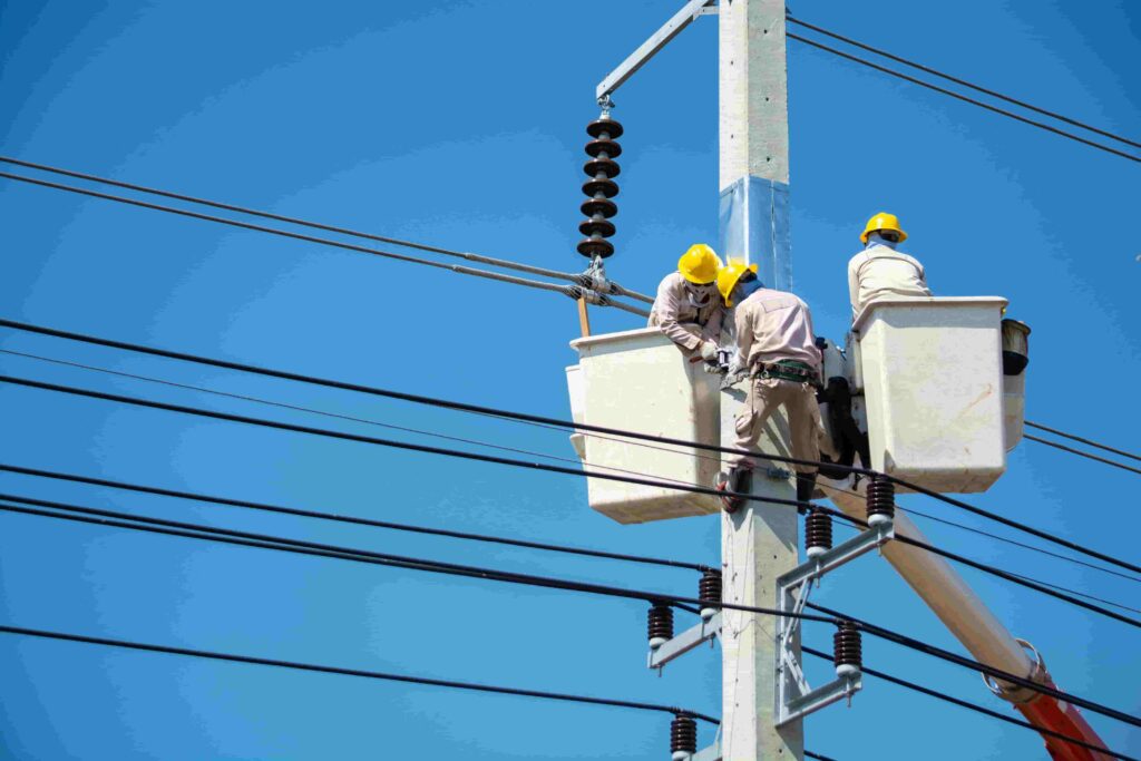 Three workers in safety gear repair electrical lines atop a utility pole using bucket lifts on a