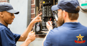 Two electricians in blue uniforms work on an open electrical panel outdoors, using tools and