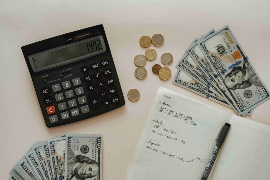 A calculator, scattered coins, stacks of 100-dollar bills, and an open notebook with handwritten