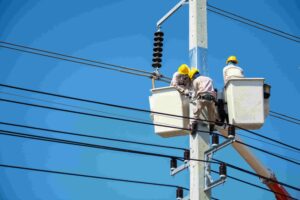 Three workers in safety gear repair electrical lines atop a utility pole using bucket lifts on a