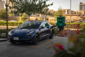 A blue Tesla car is parked at an electric vehicle charging station on a paved lot, surrounded by