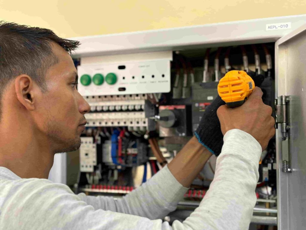 A person uses a yellow cordless drill to work on electrical components inside an open electrical