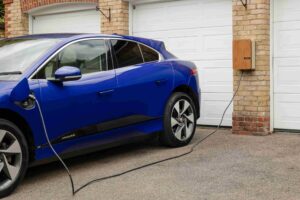 A blue Jaguar electric car is parked outside a garage and plugged into a wall-mounted charging