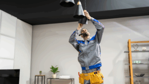 An electrician in a gray uniform and tool belt stands on a ladder, handling Lighting Design and Installation as he works on a black pendant light fixture in a modern room.