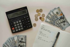 A calculator, scattered coins, stacks of 100-dollar bills, and an open notebook with handwritten