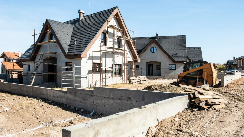 A house under construction with scaffolding, a partially built concrete wall, electrical services being installed, and a small bulldozer on a dirt lot.