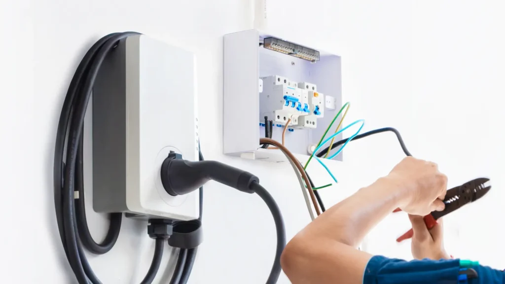 An electrician works on wiring next to an EV charger installation and an open electrical panel mounted on a white wall.